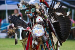 performers wearing vibrant traditional attire perform a dance at a 4th of July powwow, while spectators watch.