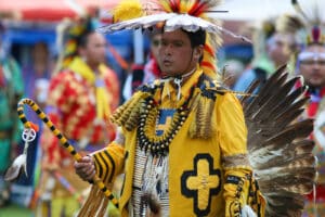 performers wearing vibrant traditional attire perform a dance at a 4th of July powwow, while spectators watch.