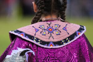 performers wearing vibrant traditional attire perform a dance at a 4th of July powwow, while spectators watch.