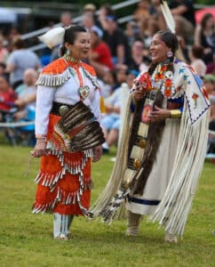 performers wearing vibrant traditional attire perform a dance at a 4th of July powwow, while spectators watch.