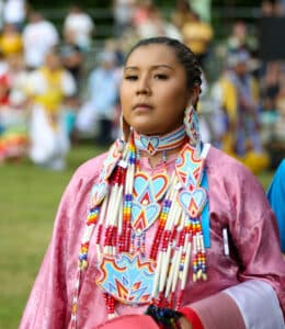 performers wearing vibrant traditional attire perform a dance at a 4th of July powwow, while spectators watch.