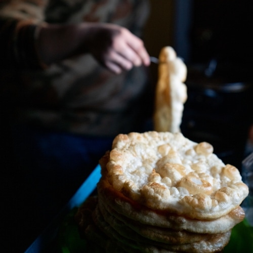 A stack of freshly fried fry bread, with a person in the background preparing food in a dimly lit kitchen. The focus is on the detailed texture of the bread's surface.