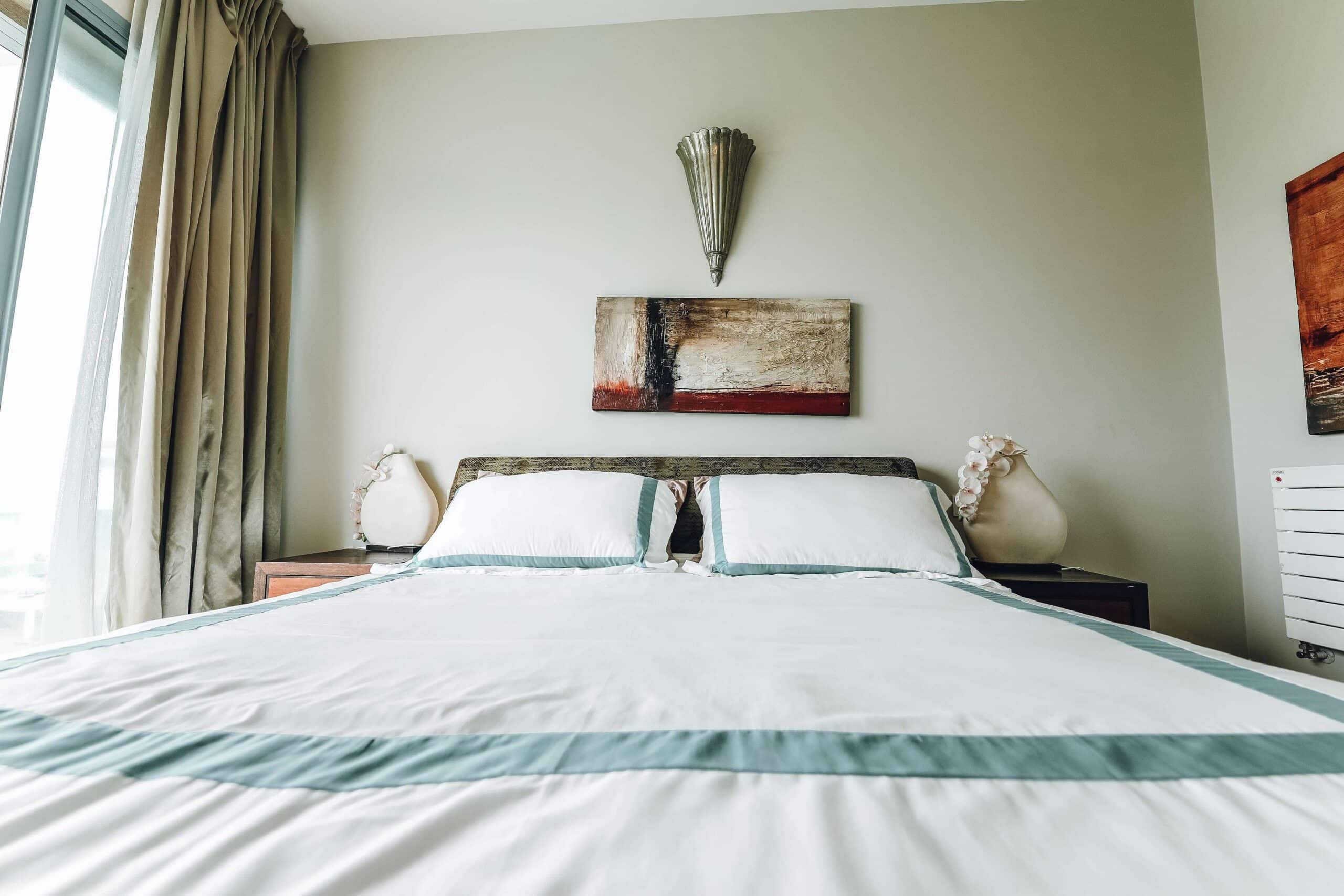 A bright, modern hotel room in Cherokee, NC featuring a large bed with white bedding and blue accents, flanked by two small lamps on bedside tables, with abstract wall art above the headboard.