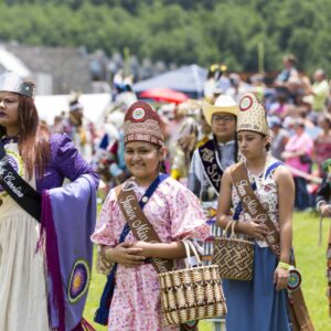 performers wearing vibrant traditional attire perform a dance at a 4th of July powwow, while spectators watch.
