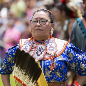 performers wearing vibrant traditional attire perform a dance at a 4th of July powwow, while spectators watch.