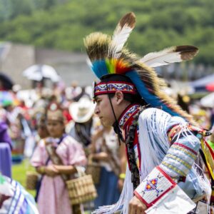 performers wearing vibrant traditional attire perform a dance at a 4th of July powwow, while spectators watch.