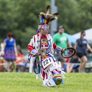 performers wearing vibrant traditional attire perform a dance at a 4th of July powwow, while spectators watch.
