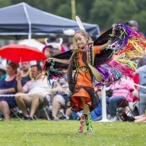 performers wearing vibrant traditional attire perform a dance at a 4th of July powwow, while spectators watch.