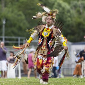 performers wearing vibrant traditional attire perform a dance at a 4th of July powwow, while spectators watch.