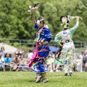 performers wearing vibrant traditional attire perform a dance at a 4th of July powwow, while spectators watch.