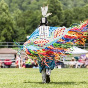 performers wearing vibrant traditional attire perform a dance at a 4th of July powwow, while spectators watch.