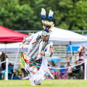 performers wearing vibrant traditional attire perform a dance at a 4th of July powwow, while spectators watch.