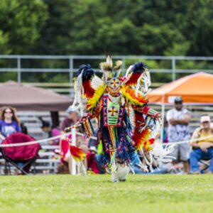 performers wearing vibrant traditional attire perform a dance at a 4th of July powwow, while spectators watch.