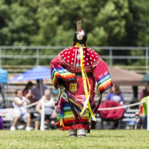performers wearing vibrant traditional attire perform a dance at a 4th of July powwow, while spectators watch.