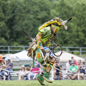 performers wearing vibrant traditional attire perform a dance at a 4th of July powwow, while spectators watch.