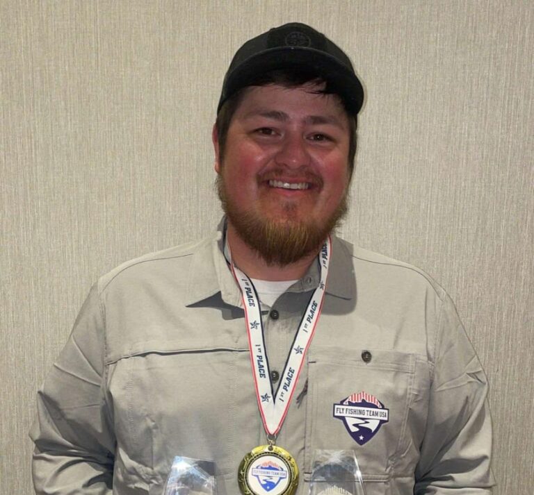 a smiling man wearing a baseball cap and a light gray shirt adorned with various patches, including a "team usa" badge. he is also wearing a medal with a ribbon around his neck.