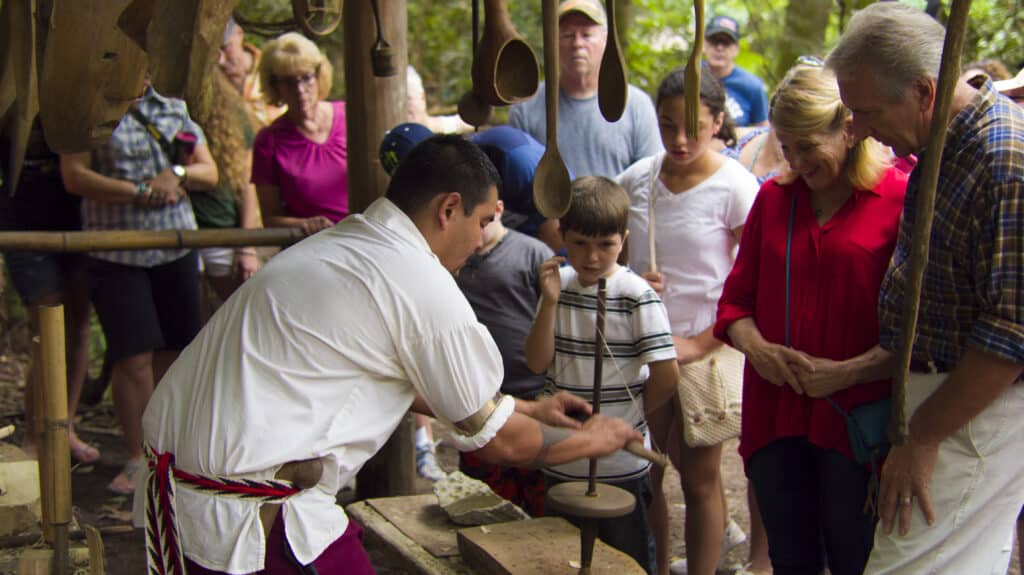 A craftsman in traditional Cherokee attire demonstrates pottery to an attentive group of visitors, including children and adults, at the Cherokee Museum in a lush outdoor setting.