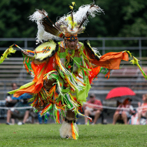 performers wearing vibrant traditional attire perform a dance at a 4th of July powwow, while spectators watch.