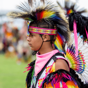 performers wearing vibrant traditional attire perform a dance at a 4th of July powwow, while spectators watch.