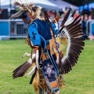 performers wearing vibrant traditional attire perform a dance at a 4th of July powwow, while spectators watch.