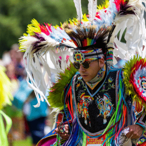 performers wearing vibrant traditional attire perform a dance at a 4th of July powwow, while spectators watch.