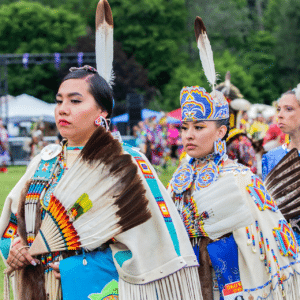 performers wearing vibrant traditional attire perform a dance at a 4th of July powwow, while spectators watch.