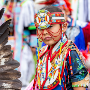 performers wearing vibrant traditional attire perform a dance at a 4th of July powwow, while spectators watch.
