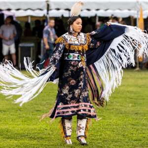 performers wearing vibrant traditional attire perform a dance at a 4th of July powwow, while spectators watch.