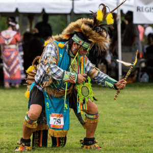 performers wearing vibrant traditional attire perform a dance at a 4th of July powwow, while spectators watch.