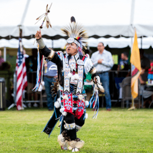 performers wearing vibrant traditional attire perform a dance at a 4th of July powwow, while spectators watch.