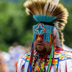 performers wearing vibrant traditional attire perform a dance at a 4th of July powwow, while spectators watch.
