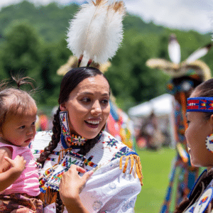 performers wearing vibrant traditional attire perform a dance at a 4th of July powwow, while spectators watch.