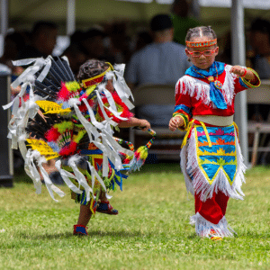 performers wearing vibrant traditional attire perform a dance at a 4th of July powwow, while spectators watch.