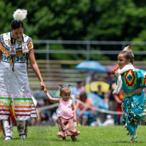 performers wearing vibrant traditional attire perform a dance at a 4th of July powwow, while spectators watch.