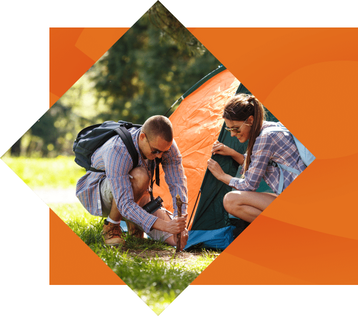 Two people, a man and a woman, are setting up a tent in a sunny, grassy park during the Cherokee Spring season. Both are wearing backpacks and casual outdoor clothing.