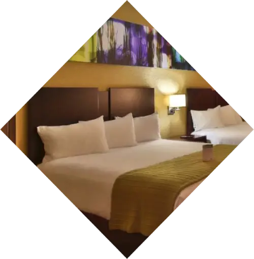 A modern hotel room featuring two double beds with white linens and a large abstract painting above the headboard. The room has soft lighting and a warm ambiance, reminiscent of Harrah's Casino Spa.