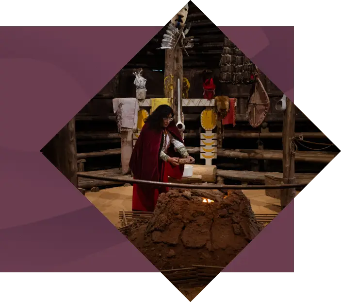 A woman in traditional attire performing a ritual at an earthen furnace, surrounded by cultural artifacts inside a dimly lit, rustic structure at the Oconaluftee Indian Village.