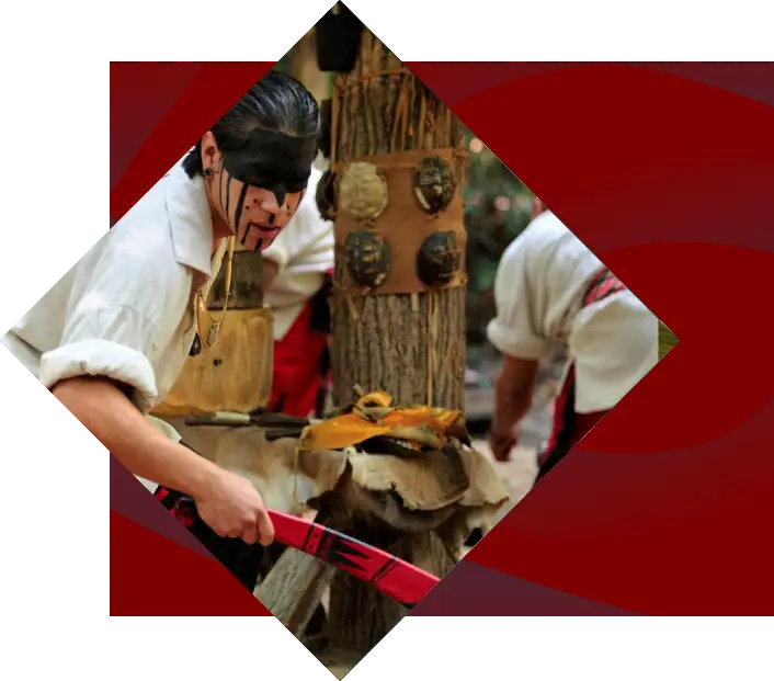 A man wearing traditional clothing and a black eye mask chops wood at Oconaluftee Indian Village, with another participant partially visible in the background. The setting includes cultural decorations.