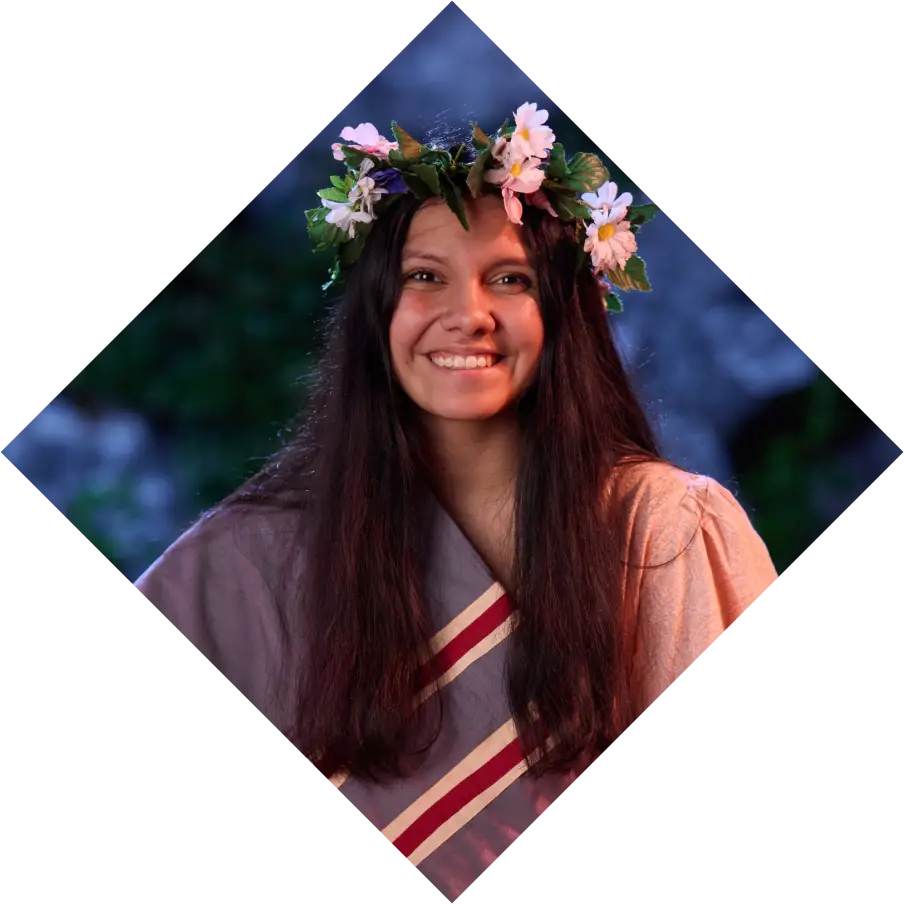 A woman with long hair wearing a flower crown smiles warmly in a softly lit evening setting, framed within a diamond-shaped border, capturing the essence of an "Unto These Hills" performer.